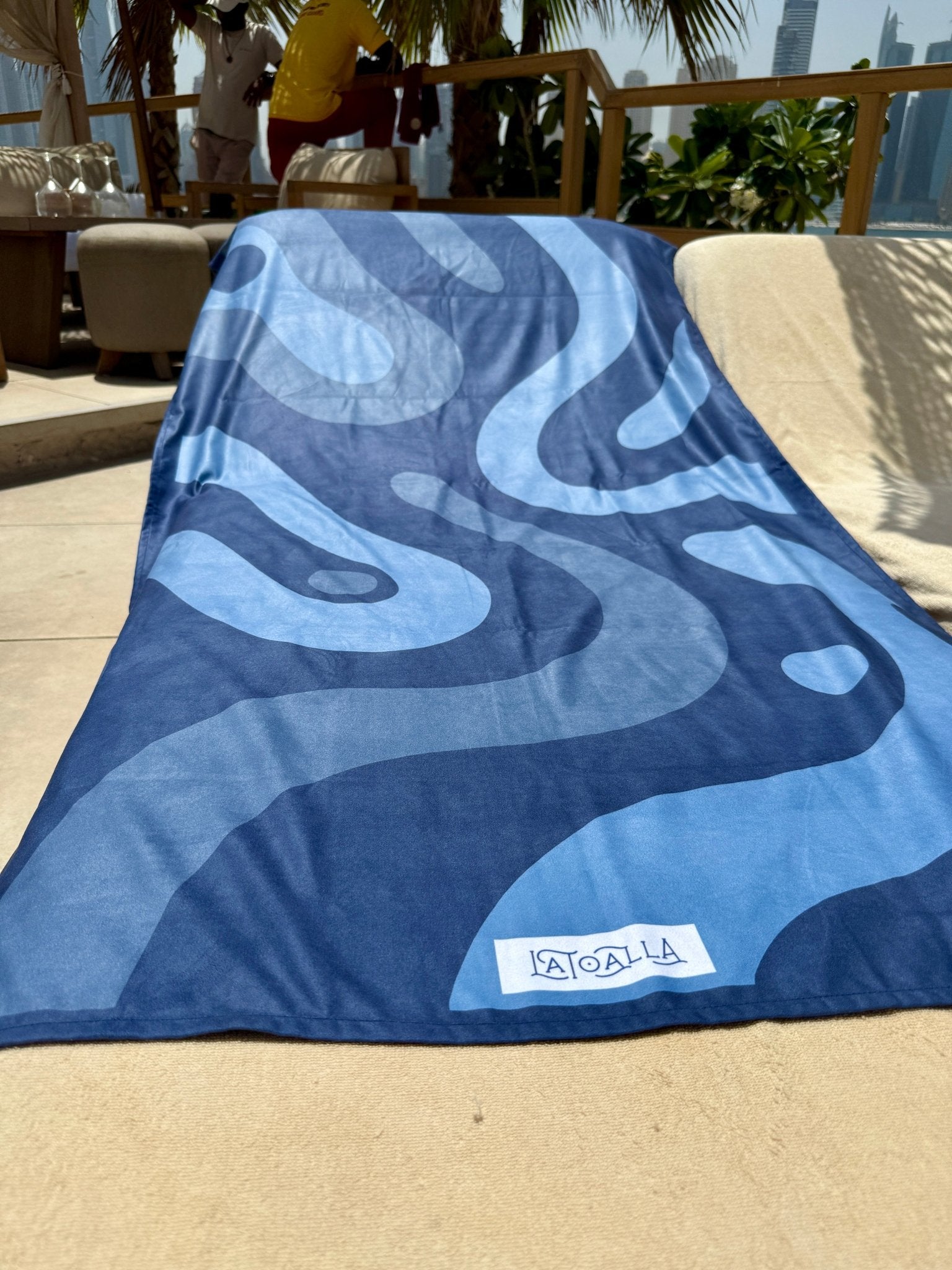 Blue towel with a pattern on a sandy surface, likely at a beach or poolside.