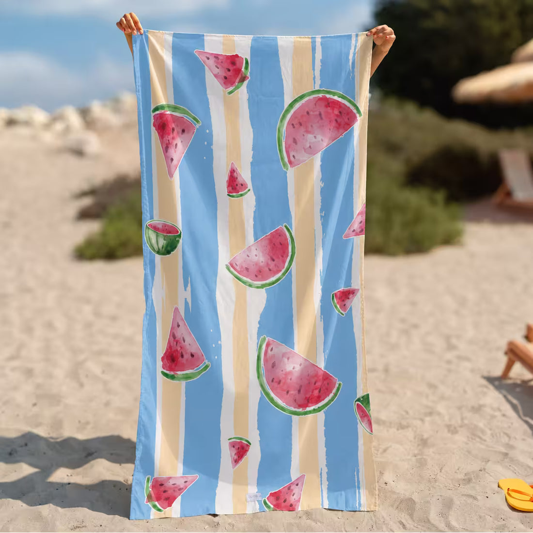 A person holds up a large beach towel from la Toalla on a sandy beach. The towel features a light blue background with vertical cream stripes and watermelon slice illustrations in pink, red, and green.