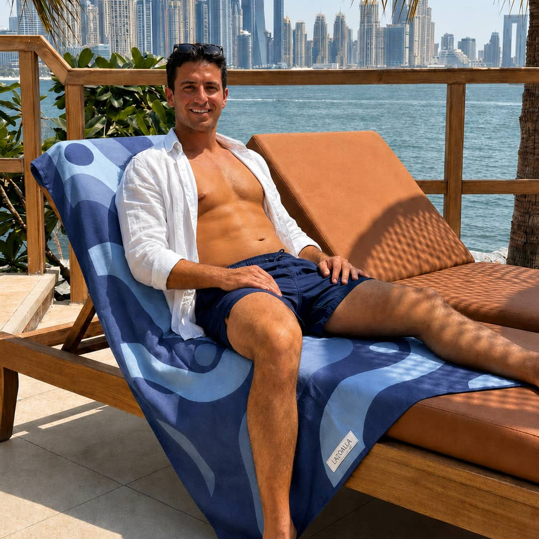 A man relaxes on a brown leather chaise lounge with a blue patterned towel, set on a patio with a waterfront and a city skyline in the background.